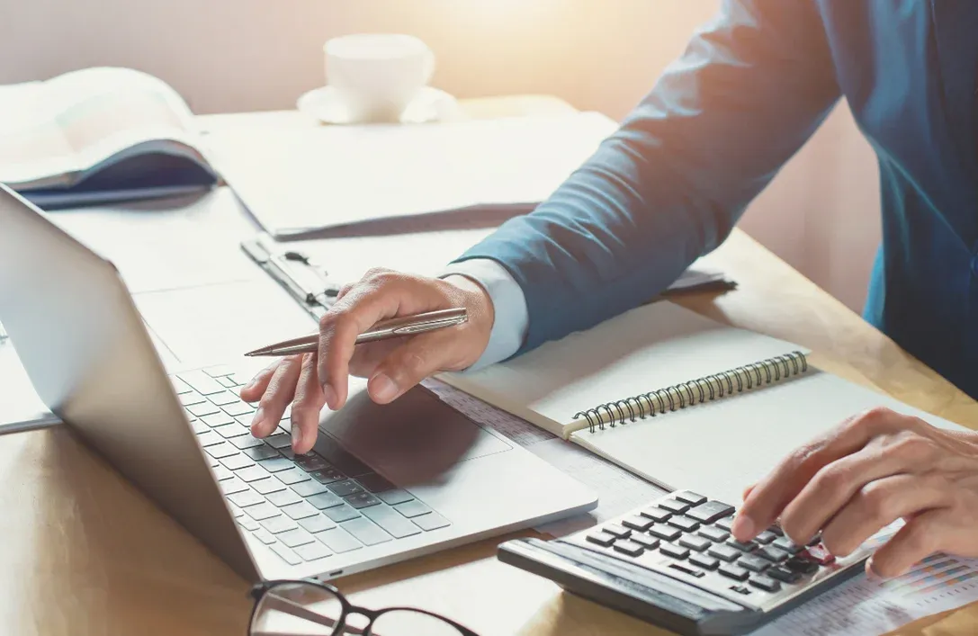 A business professional using a laptop and calculator while working on financial documents, representing efficient financial management for fleet operations. This image illustrates the process of calculating fuel tax credits (FTC), with notebooks and spreadsheets visible in the background. Geosecure partners with experts such as KPMG and Ryan to offer data-driven, compliant solutions for optimising fuel tax credits in the transport and logistics sector. The focus is on utilising advanced data science and telematics to maximise credits and ensure accurate reporting for fleets across various industries.