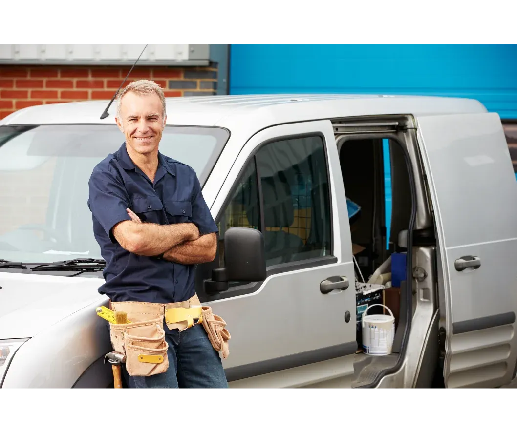 A male tradesperson in a work shirt stands confidently in front of a silver service van, tools ready, at a job site.