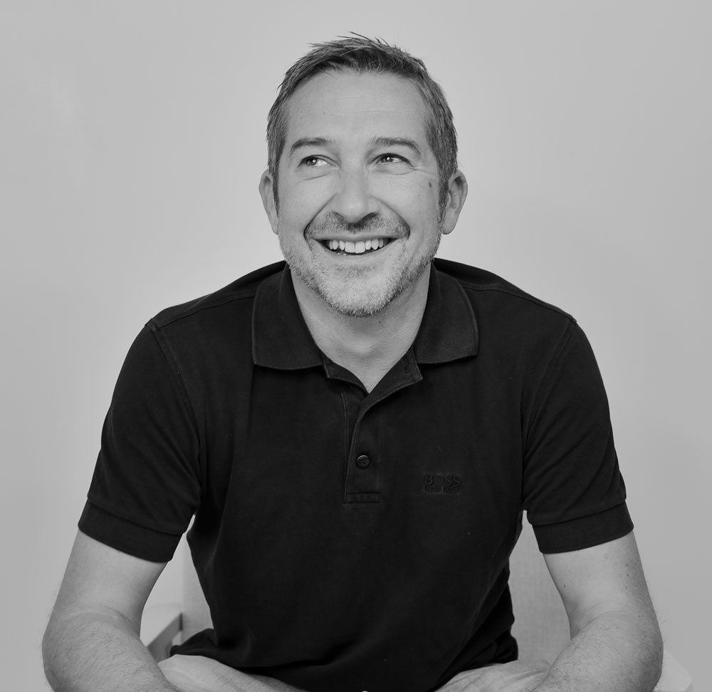 A black-and-white portrait of Andrew Hudson, Director of Geosecure, wearing a short-sleeve polo shirt with a subtle ''BOSS'' logo on the chest. Andrew is seated, smiling warmly, looking slightly off to the side, exuding a confident yet approachable demeanour. His relaxed posture, with arms resting on his legs, gives a casual yet professional vibe. The plain, neutral background places the focus entirely on him. Andrew is known for his extensive 20+ years of experience in advanced software solutions, particularly in the transportation industry, and his leadership style is characterised by openness, honesty, and integrity. 